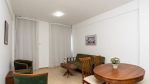 Uma área de refeições com mesa e cadeiras de madeira, uma pequena mesa redonda com uma garrafa, um sofá verde e um vaso de planta.