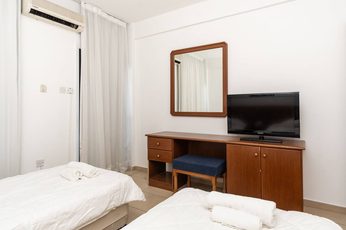 Habitación de hotel con dos camas, un escritorio de madera con televisión, un espejo y una ventana con cortinas blancas.