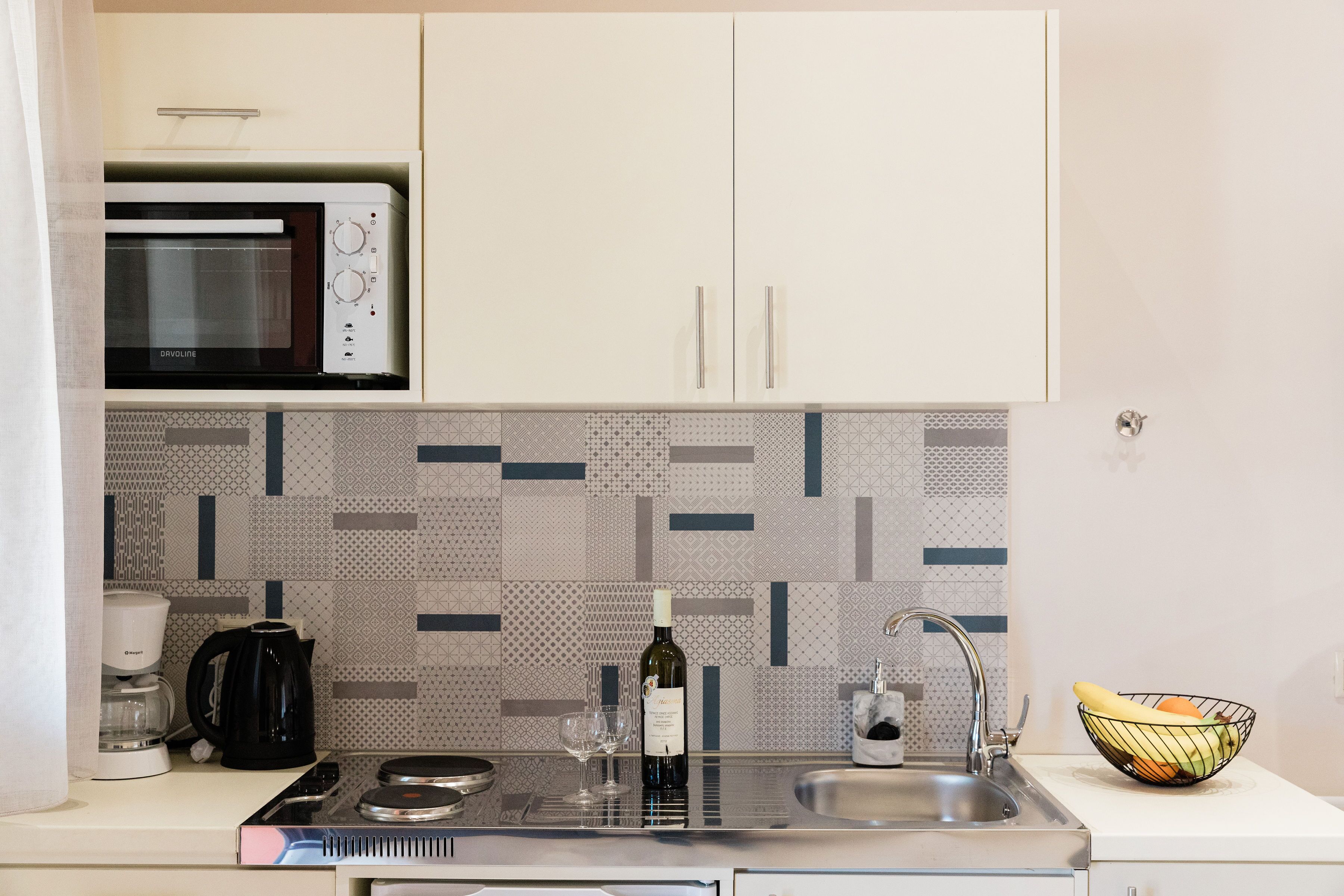 A kitchen with a microwave, coffee maker, wine bottle, glasses, and a fruit bowl.