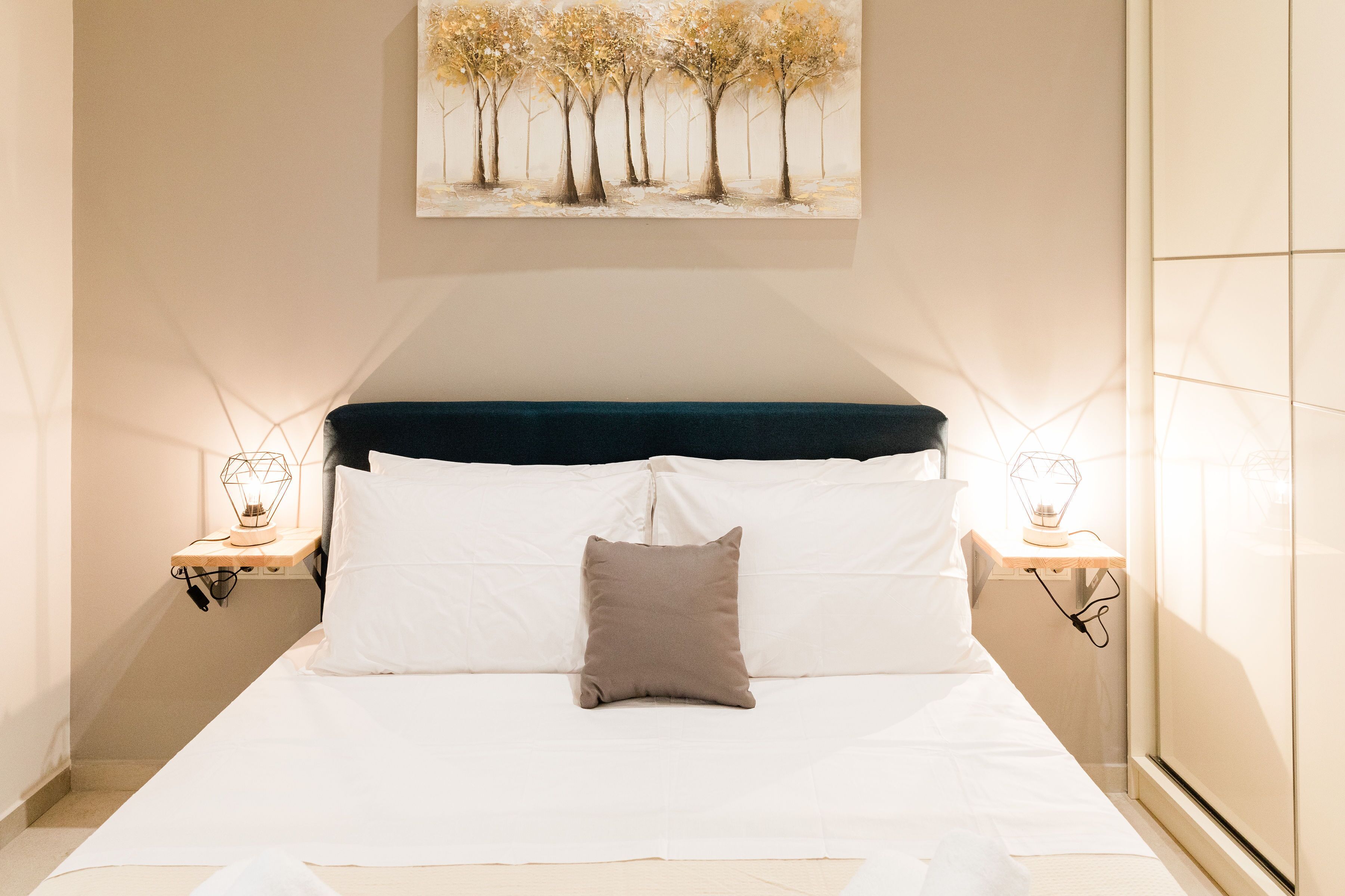 Een net opgemaakt bed met witte lakens en een enkel bruin kussen, geflankeerd door twee nachtkastjes met geometrische lampen, in een kamer met een schuine kap en een schilderij van bomen boven het bed.