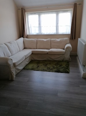 A corner living room with a beige sofa, a green rug, and a window with curtains. - Apartment near Heathrow Airport & Central London (Ickenham)