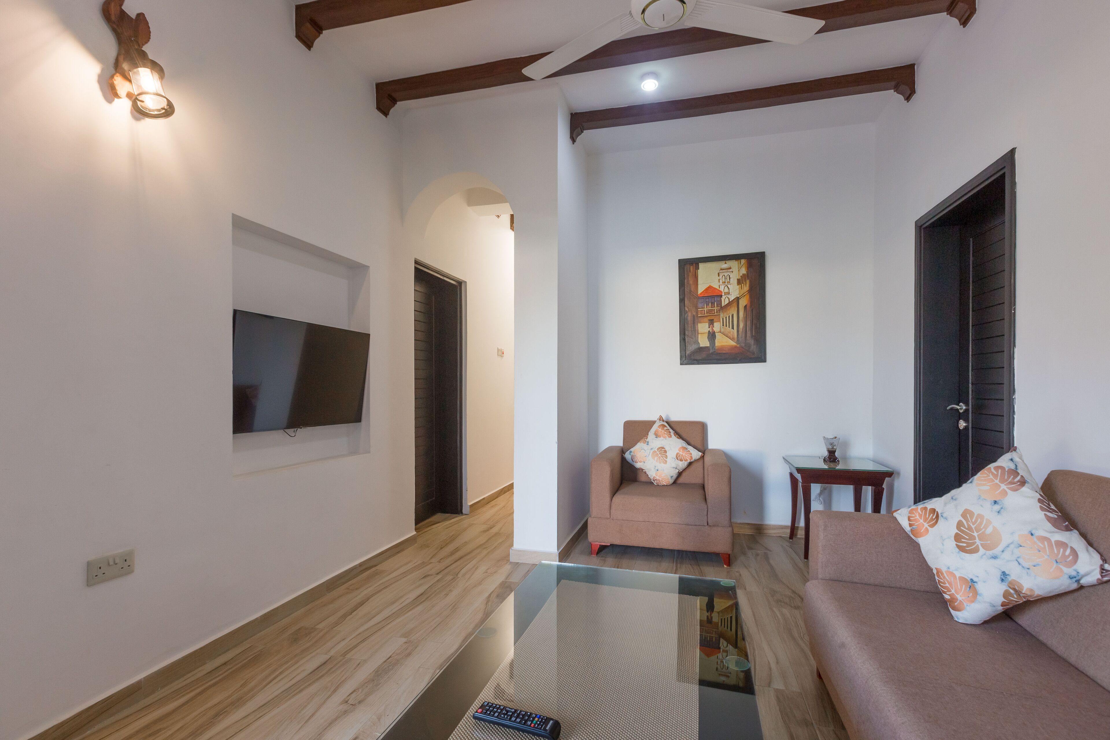 A living room with a sofa, a chair, a glass coffee table, a television mounted on the wall, a painting, and a door.