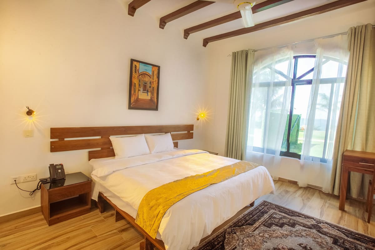A bedroom with a wooden bed, a bedside table with a phone, a painting on the wall, and a window with curtains.