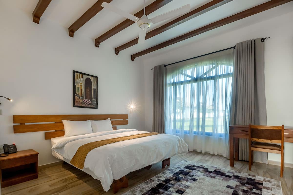 A bedroom with a wooden bed, a desk, a chair, a large window with curtains, and a ceiling fan.