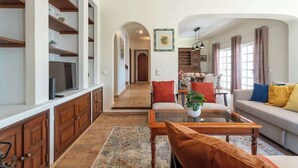 Un vaste salon comprenant un canapé, une table basse et un téléviseur posé sur un meuble en bois. On aperçoit à l’arrière-plan un coin repas avec des chaises et une table.