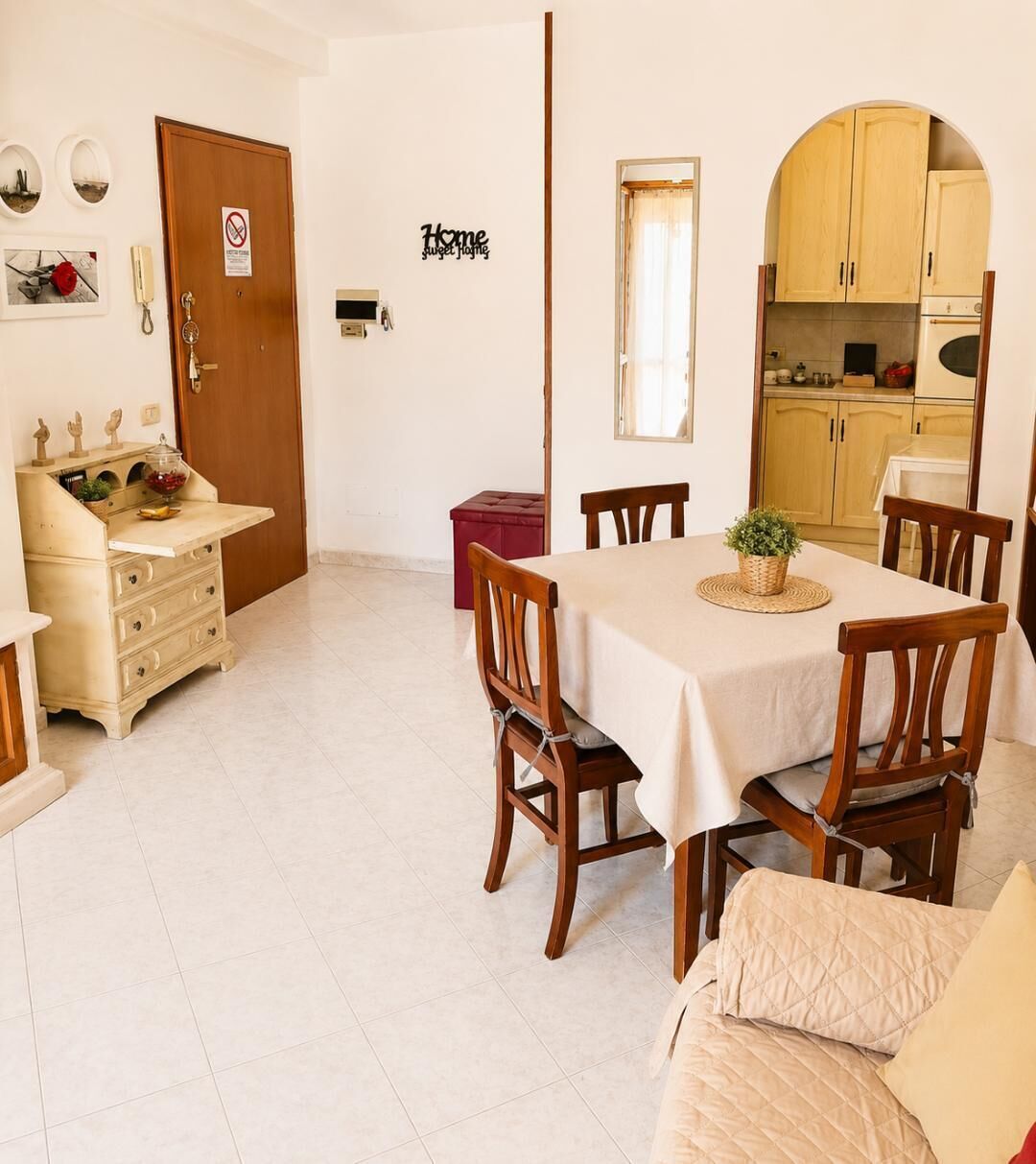 Un comedor con una mesa dispuesta para cuatro, sillas de madera, un reposapiés rojo, un sofá color crema, y una cocina visible a través de un arco.