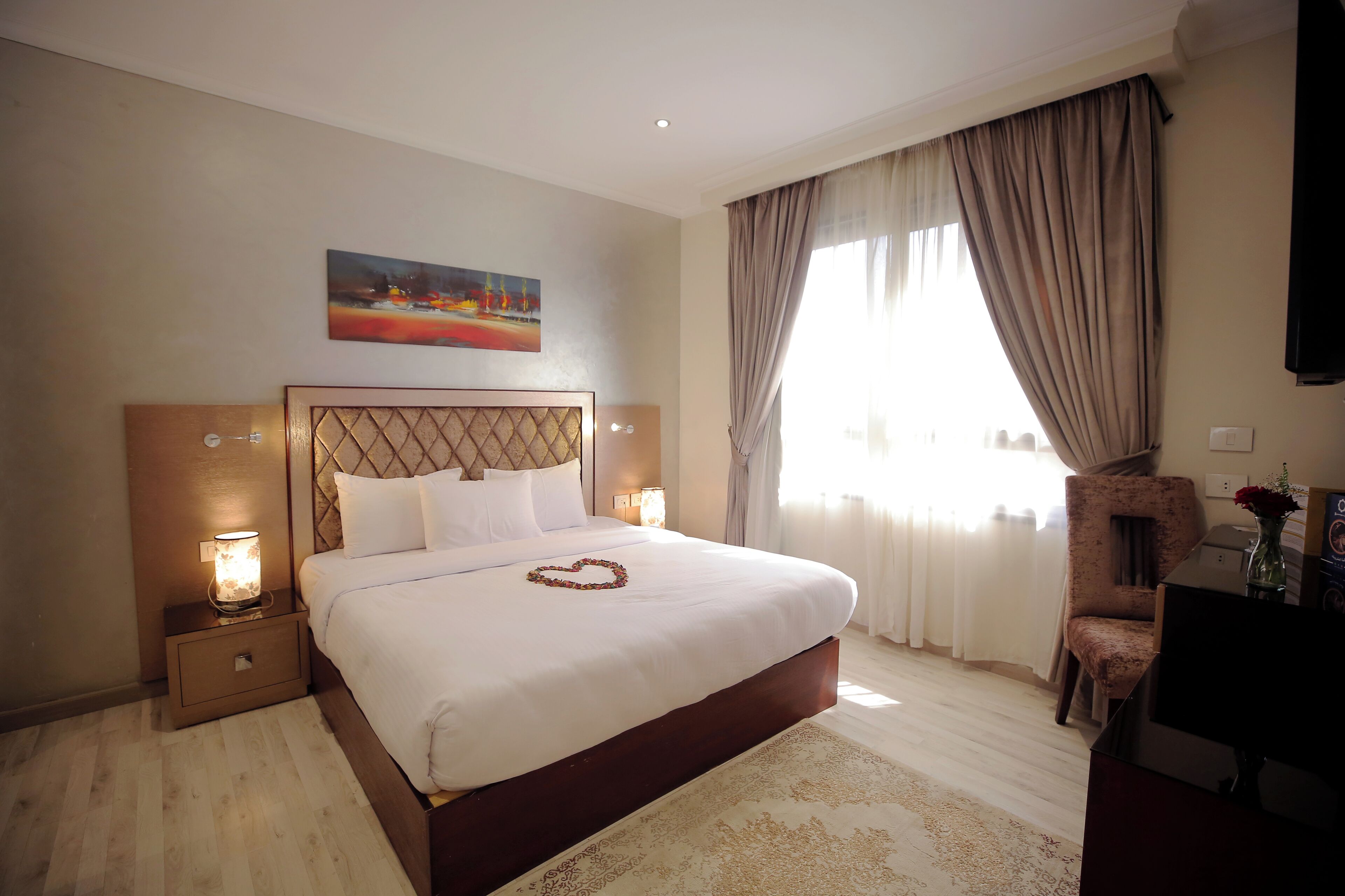 A hotel room with a large bed, a nightstand with a lamp, a chair, a desk, and a painting on the wall.