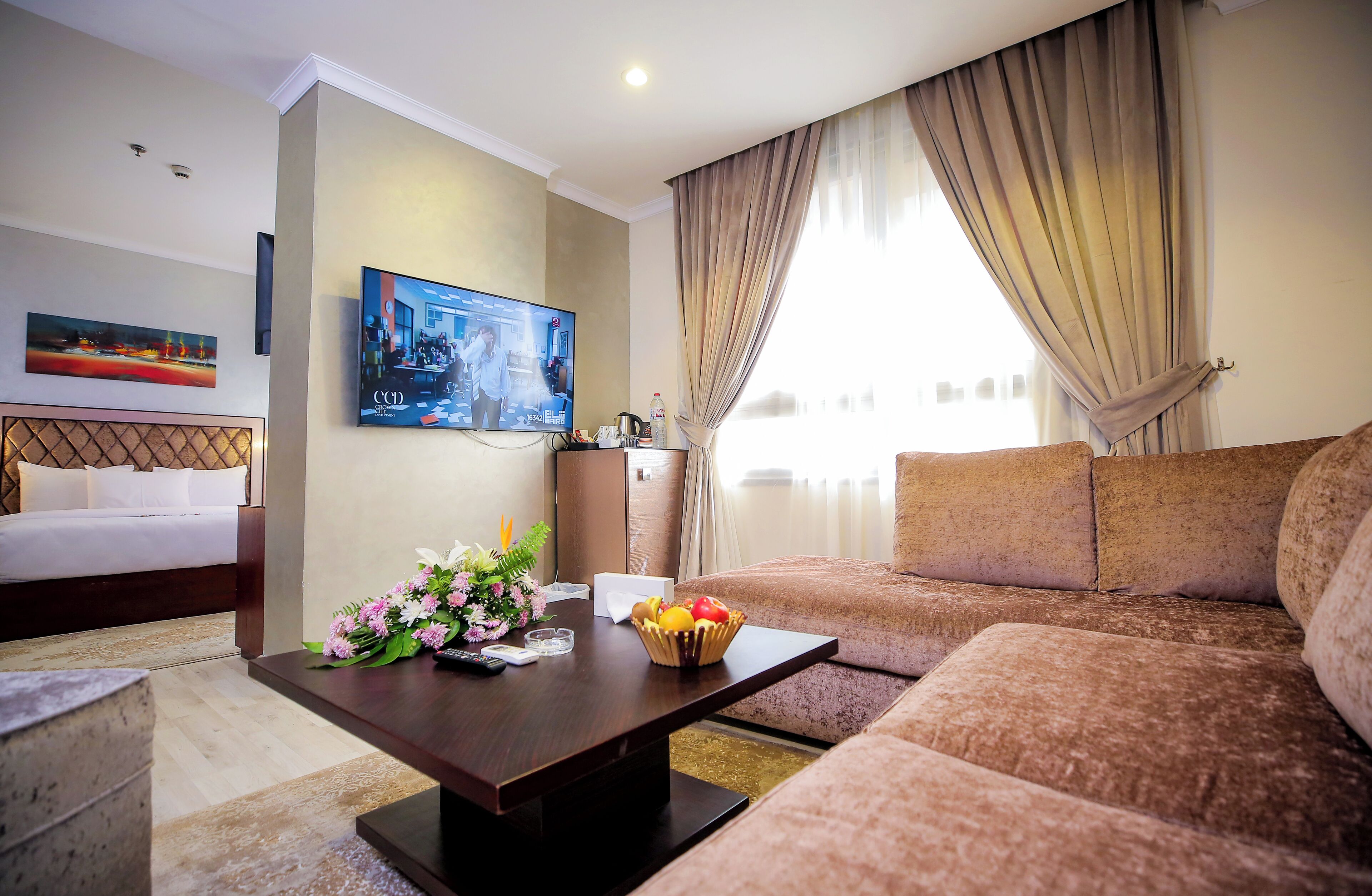 A hotel room with a bed, a sofa, a coffee table with flowers and fruit, a TV, and a window with curtains.