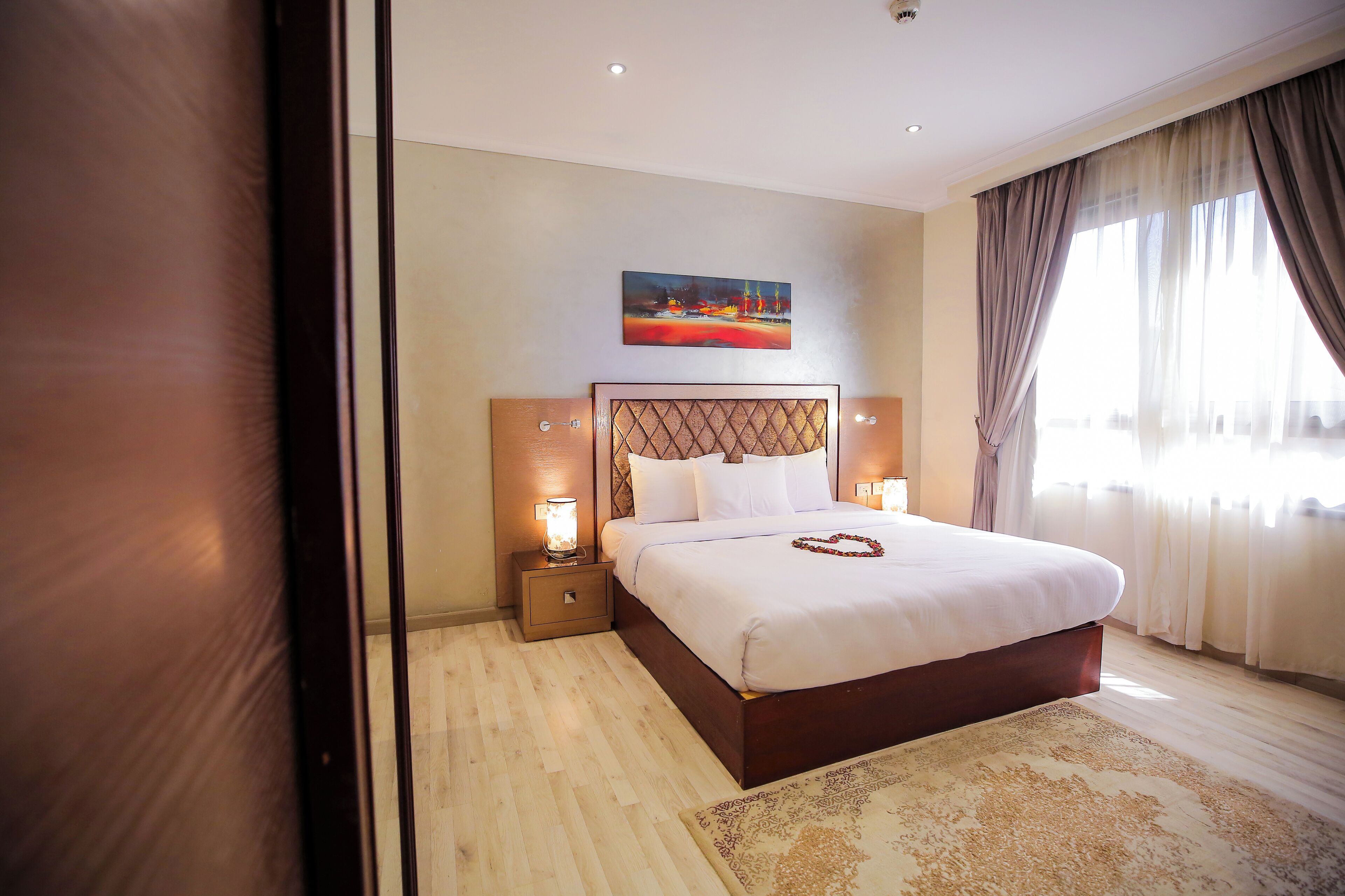 A neatly made bed with a headboard, two bedside lamps, and a painting above it.