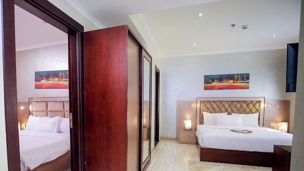 A hotel room with a large bed, a smaller bed, and a walk-in closet. There are two nightstands with lamps and a painting on the wall. - Cairo Taj Hotels & Suites - Dokki (Giza)