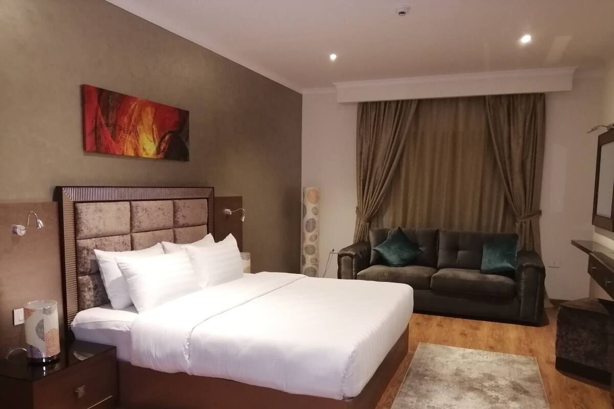 A bedroom with a bed, a nightstand, a dresser, a dark leather sofa, a large painting, and a window with curtains.