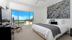 A bedroom with a large window, a bed with white and gray bedding, a black and white painting, and a view of a beach and buildings. - Sapphire on Wilson, Gerringong (Gerringong)