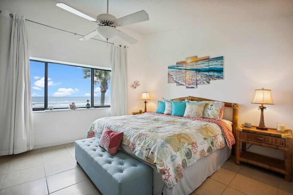 A bedroom with a large bed, a ceiling fan, a window with a view of the beach, and two bedside tables with lamps.