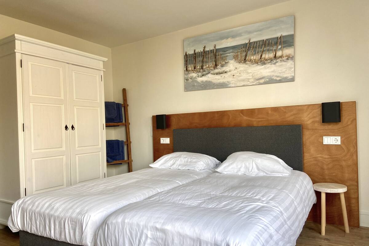 Une chambre à coucher avec un grand lit, une tête de lit en bois, un tableau au mur, une armoire et un petit tabouret.