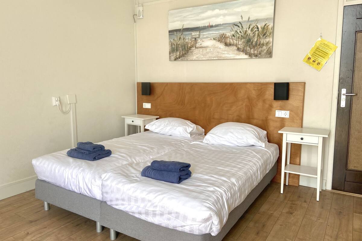 Un lit bien fait, avec du linge de lit blanc et des serviettes bleues, une tête de lit en bois, des tables de chevet avec des lampes et un tableau sur le thème de la plage accroché au mur.