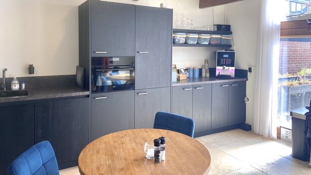 A modern kitchen with dark cabinetry, a built-in oven, and a coffee machine. There is a dining table with chairs and a window providing a view of the outside. - Bed and Ontbijt Haddock (Almere)