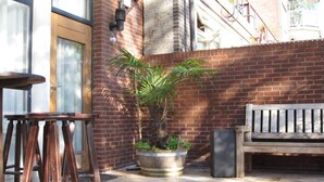 A patio area with a brick wall, a wooden bench, a metal planter with a tree, and a wooden stool. - Bed and Ontbijt Haddock (Almere)