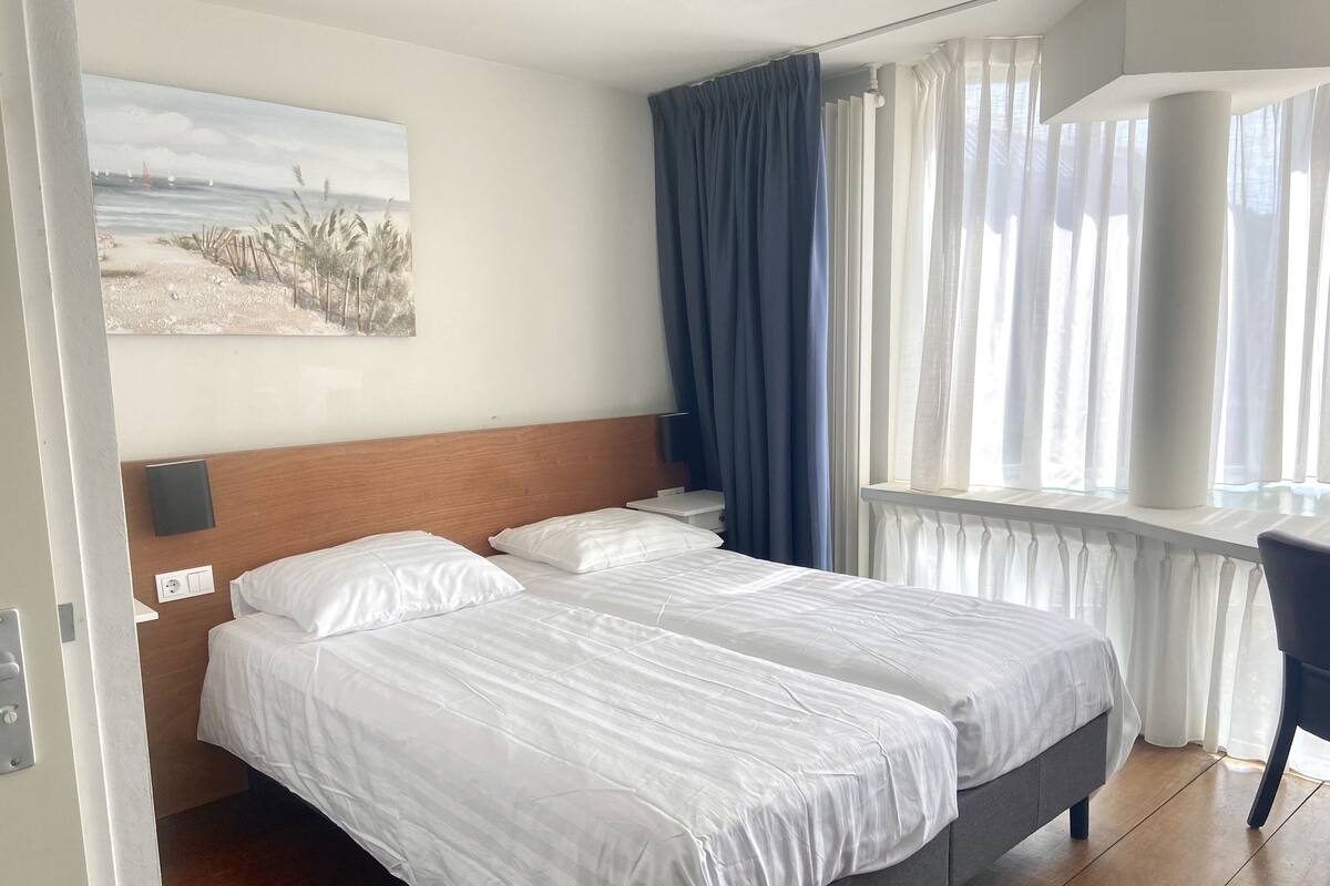 Une chambre d’hôtel avec deux lits, un tableau représentant une plage, un bureau et une fenêtre avec des rideaux.