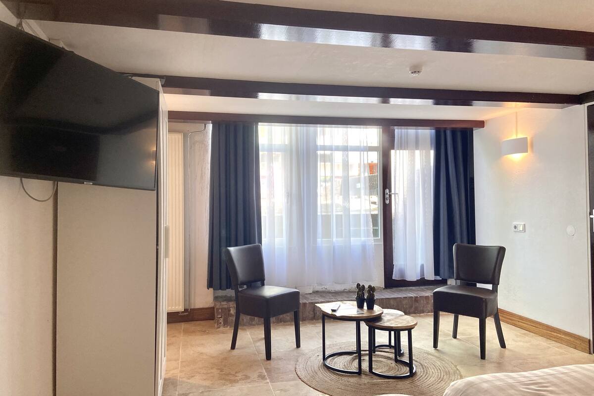Une chambre d’hôtel comprenant un lit, deux chaises, une petite table, un téléviseur fixé au mur et une fenêtre avec des rideaux.