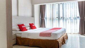 A hotel room with a large bed, two red pillows, a white headboard, and a view of a building through sheer curtains. - OYO 3811 Best Inn Hotel (Jakarta)