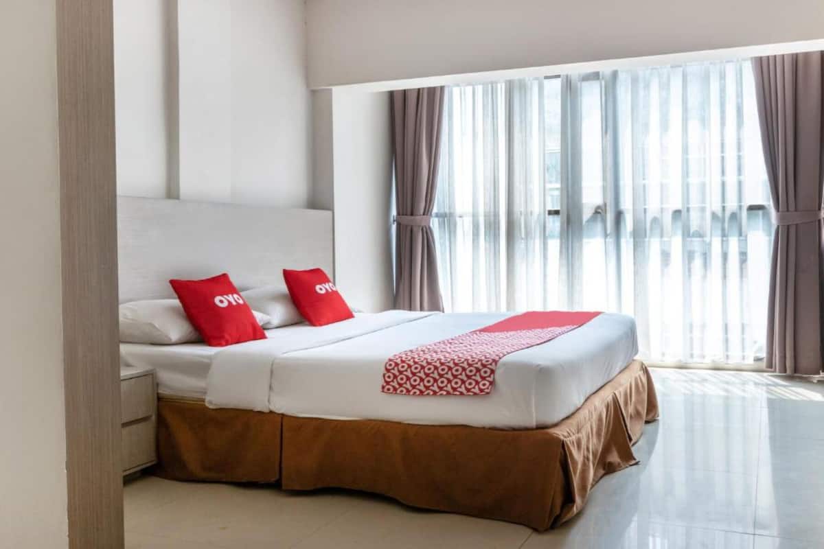 A hotel room with a large bed, two red pillows, a white headboard, and a view of a building through sheer curtains.
