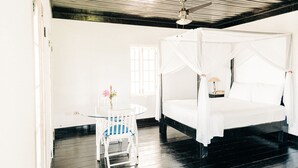 A room with a four-poster bed, a round table with a chair, a ceiling fan, and a window with white curtains.