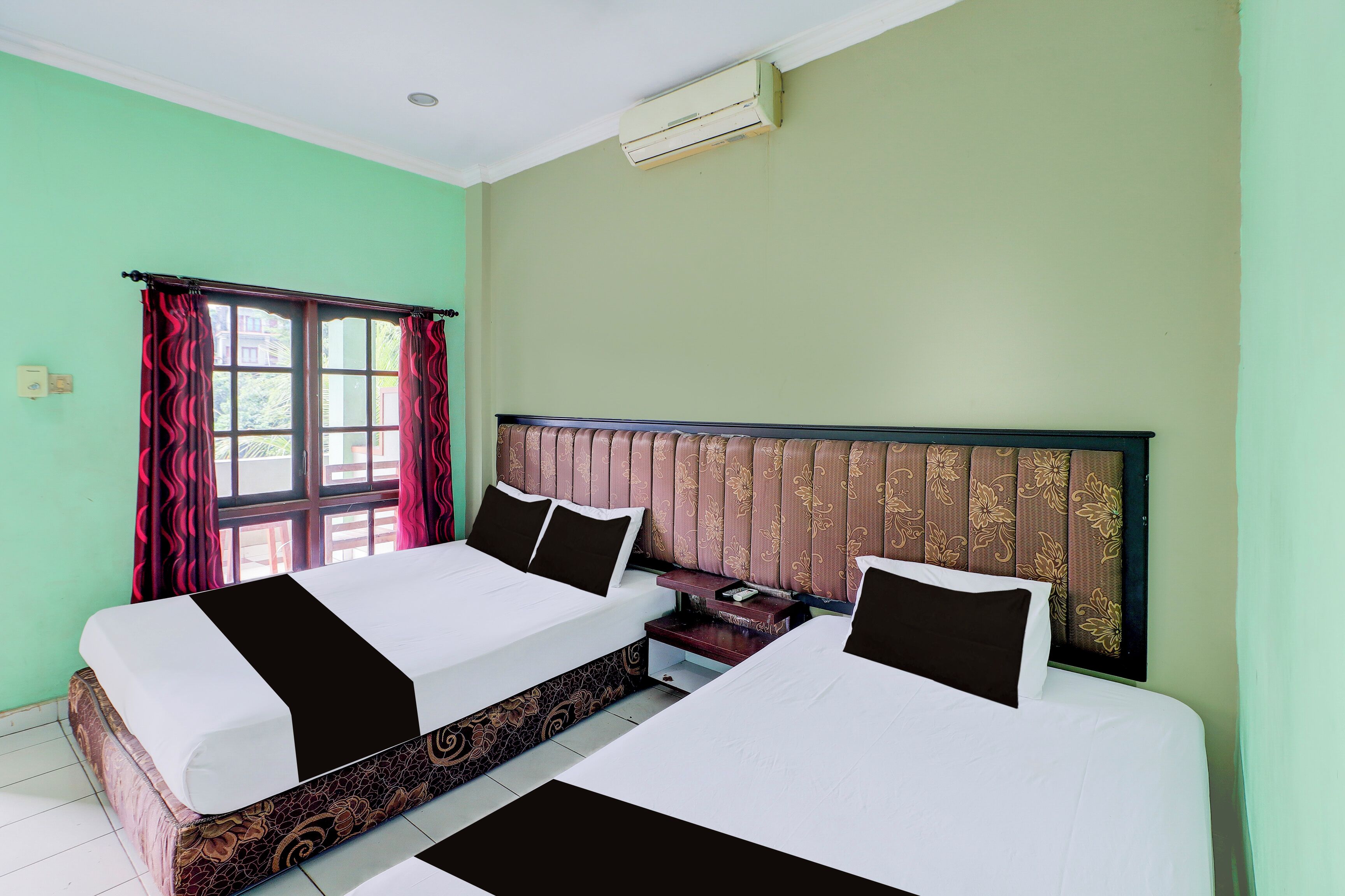 A hotel room with two beds, a window with curtains, a headboard with a floral pattern, and an air conditioning unit on the wall.