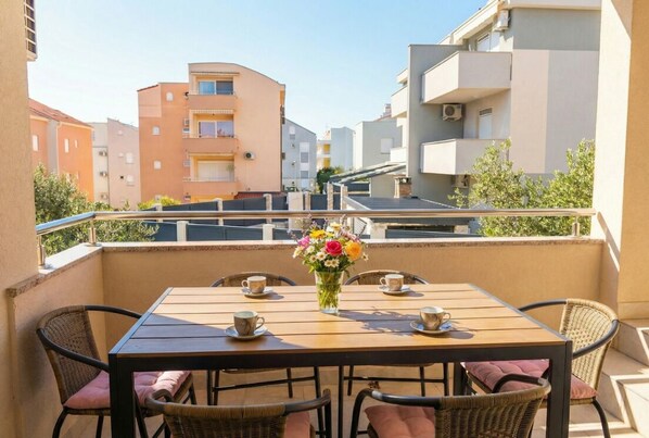A balcony with a wooden table and wicker chairs, a vase with flowers, and a view of surrounding buildings.