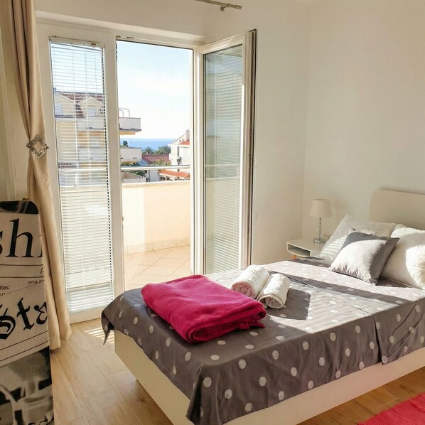 Quarto com cama, travesseiros, mesa de cabeceira e vista para o mar através de uma porta de correr de vidro.