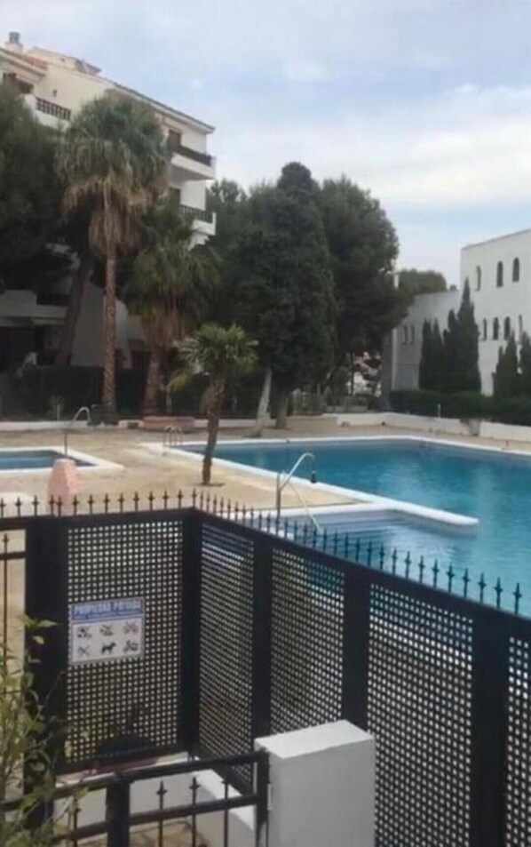 A swimming pool with a black fence, surrounded by trees and buildings. - Manila Playa Pp2 PET (Alcocéber)