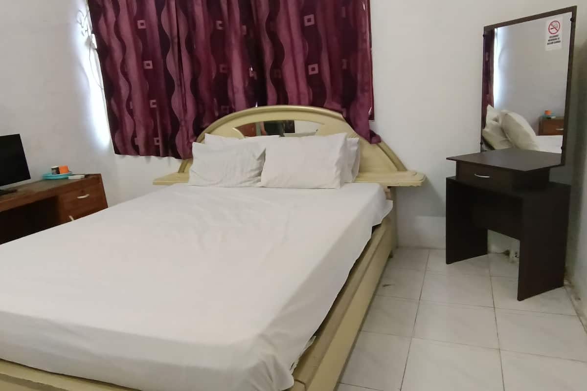 Cama com roupa de cama branca, cabeceira de madeira, mesa de cabeceira com televisão, espelho e outra mesa de cabeceira escura.