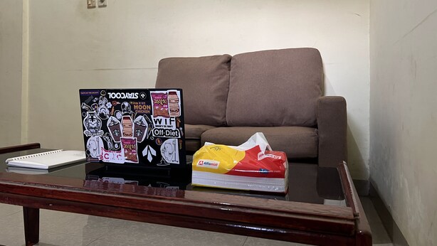 A room with a brown sofa, a glass coffee table with a notebook and a sticker-covered laptop, and a package of tissues. - SPOT ON 92338 Karin Kost (Karang Intan)