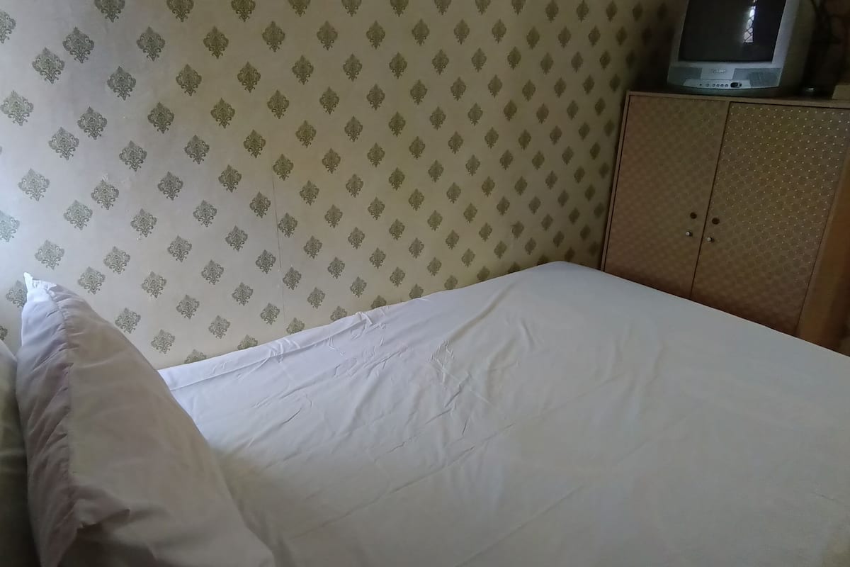 Cama de solteiro com roupa de cama branca em quarto com papel de parede estampado e televisão sobre um móvel.