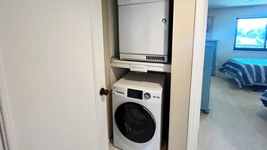 Un lavadero con lavadora y secadora de carga frontal, una puerta y vistas a un dormitorio con cama y ventana.