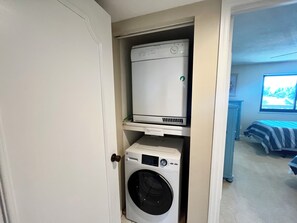 A laundry room with a front-loading washer and dryer, a door, and a view into a bedroom with a bed and window. - BT 3942 Pond View Condo-Welcome to Paradise (Fort Pierce)