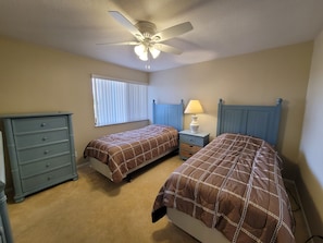 Une chambre avec deux lits, une commode, une table de chevet avec une lampe, un ventilateur de plafond et une fenêtre avec des stores.