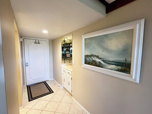 A hallway with a white door, a tiled floor, a framed painting of a seascape, and a small section of a kitchen with cabinets. - BT 3942 Pond View Condo-Welcome to Paradise (Fort Pierce)