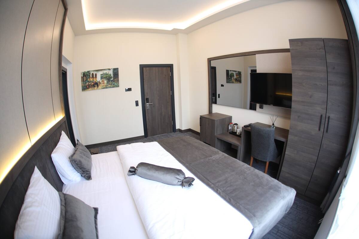 A modern hotel room with a large bed, a desk, a chair, a wardrobe, and a TV.