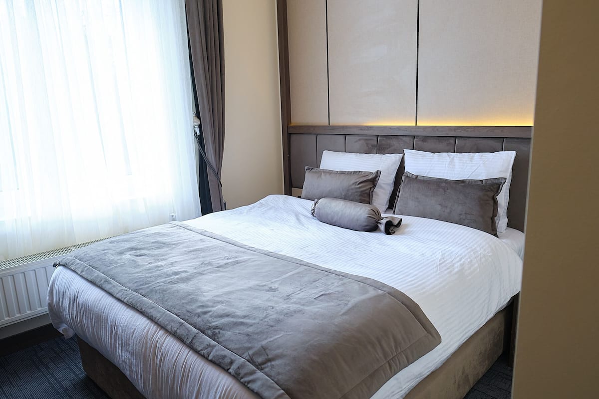 A neatly made bed with a grey duvet and pillows, a wooden headboard, and a window with sheer curtains.