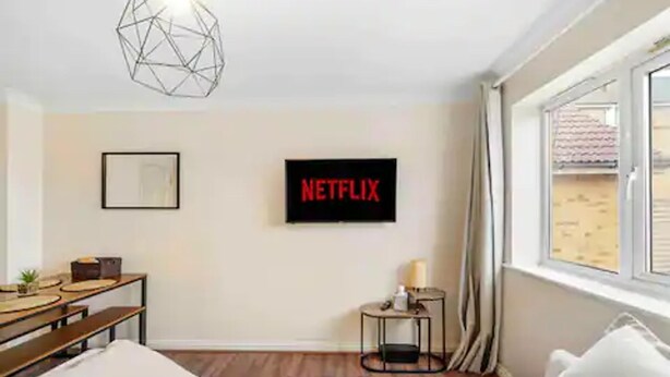A modern hotel room with a bed, a desk with a computer, a small table with a lamp, a wall-mounted TV displaying 'NETFLIX', and a window with curtains. - 4 bed Townhouse- Free Parking (London)
