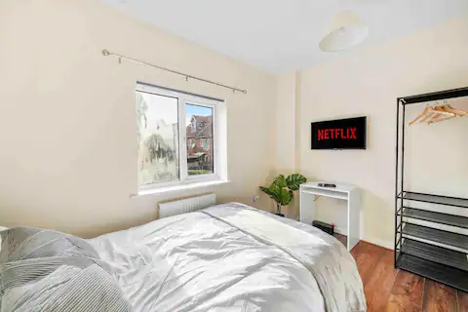 Ein Schlafzimmer mit Bett, Fenster, Pflanze, Schreibtisch und einem an der Wand befestigten Fernseher.