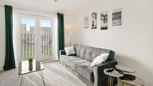 Ett modernt vardagsrum med en grå soffa, ett litet runt bord och en golvlampa. Det finns gröna gardiner på skjutdörren i glas, och tre inramade tavlor på väggen.