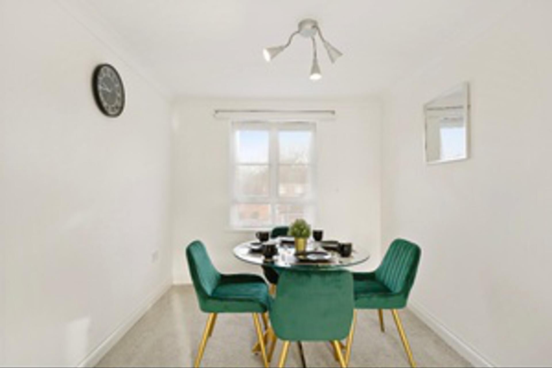 A dining area with a round table, two green chairs, a ceiling light fixture, a window, and a wall clock.