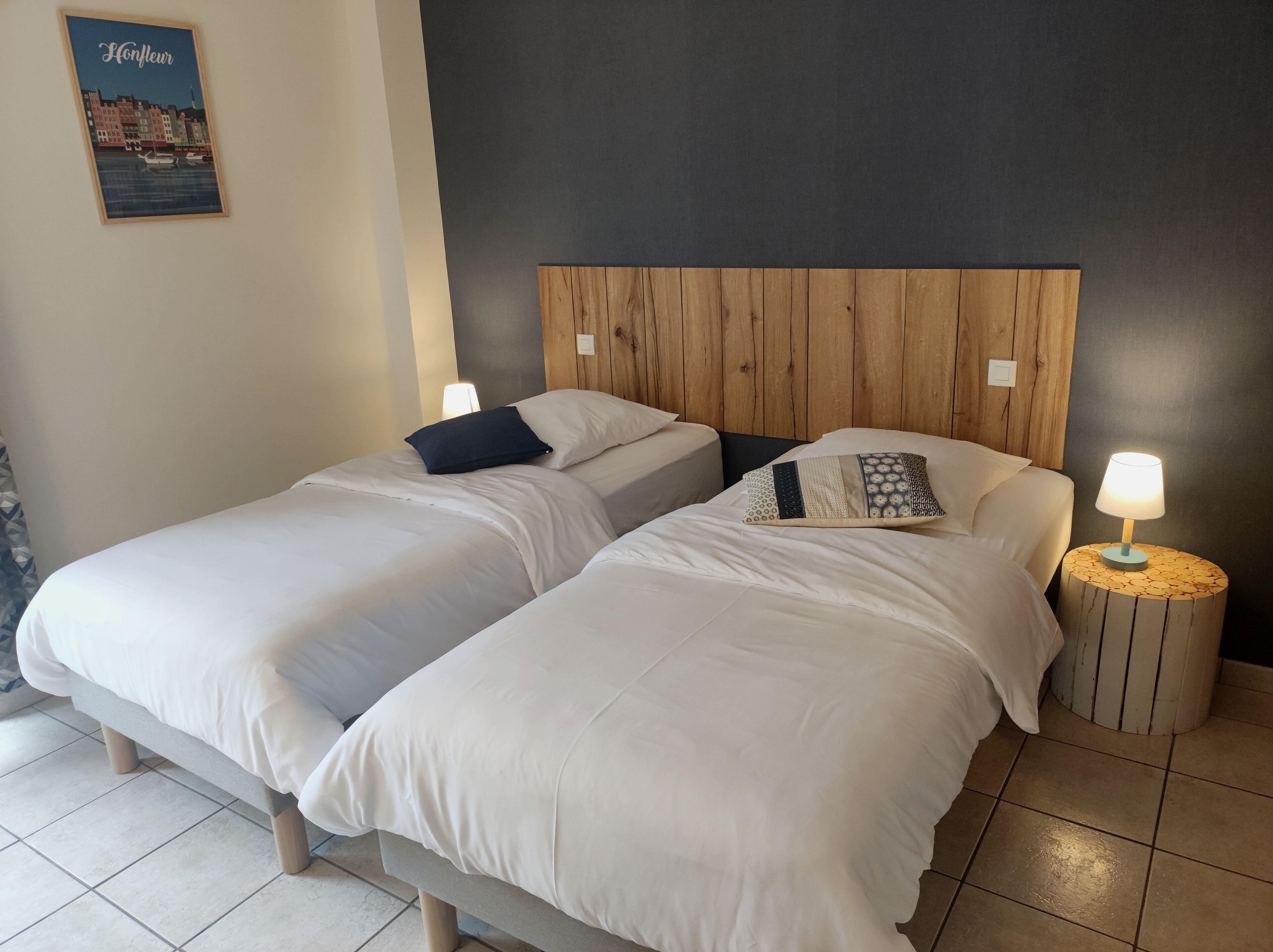 Une chambre d’hôtel avec deux lits simples, une tête de lit en bois, une table de chevet avec une lampe et un tableau encadré au mur.