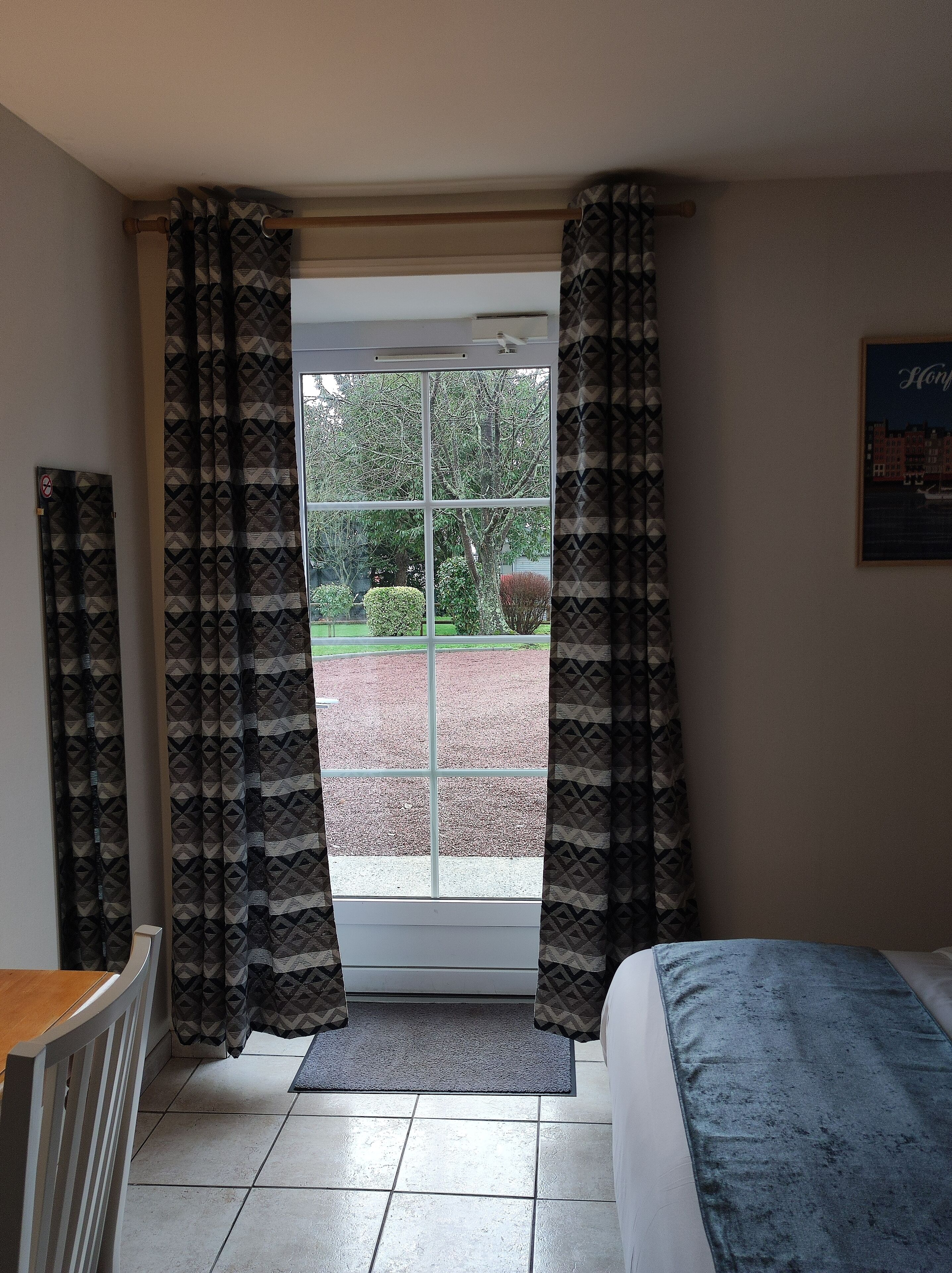 A room with a bed, a window with patterned curtains, a radiator, and a door leading outside.