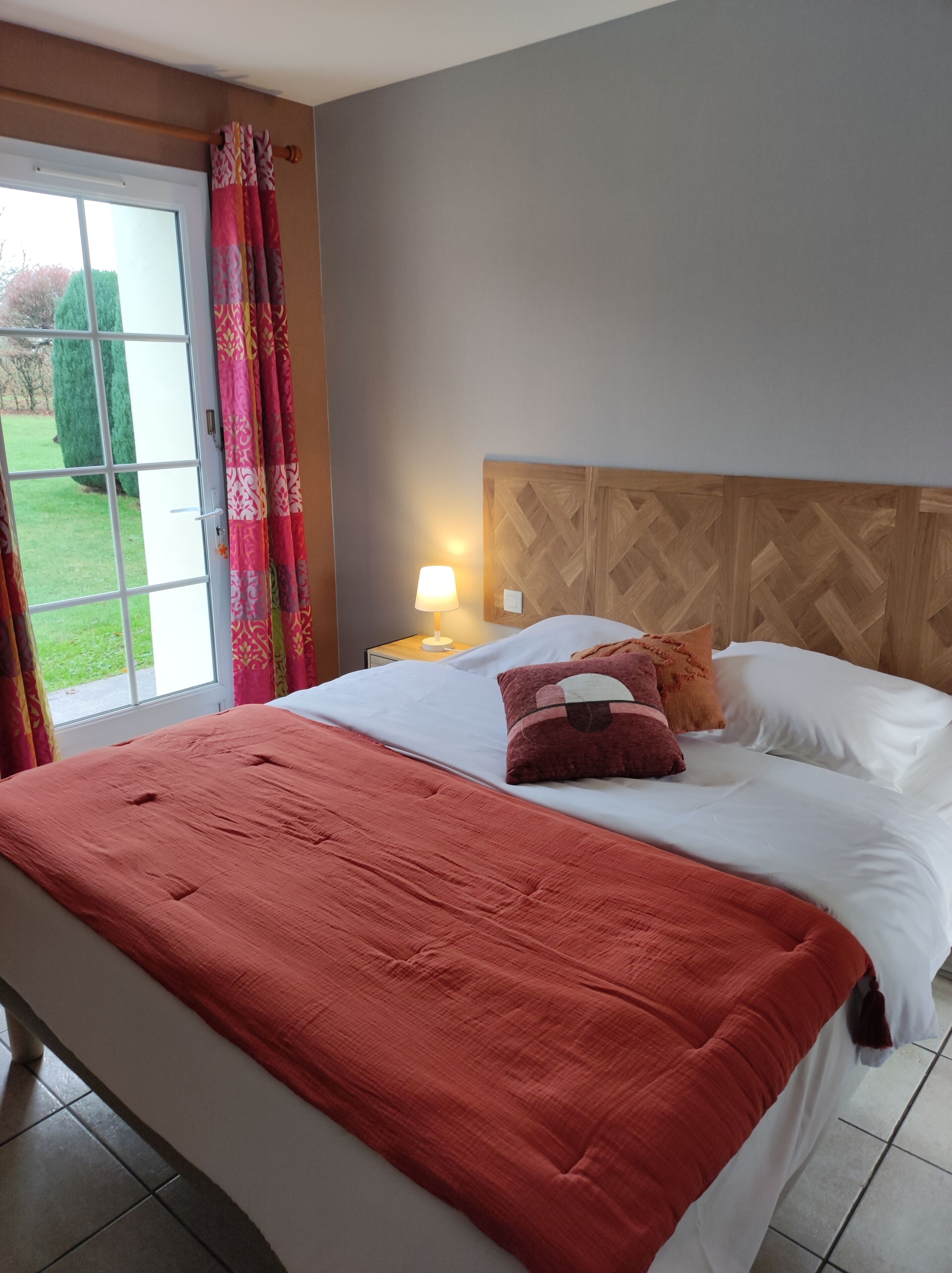 A hotel room with a bed, a nightstand, a lamp, and a view of the outdoors.