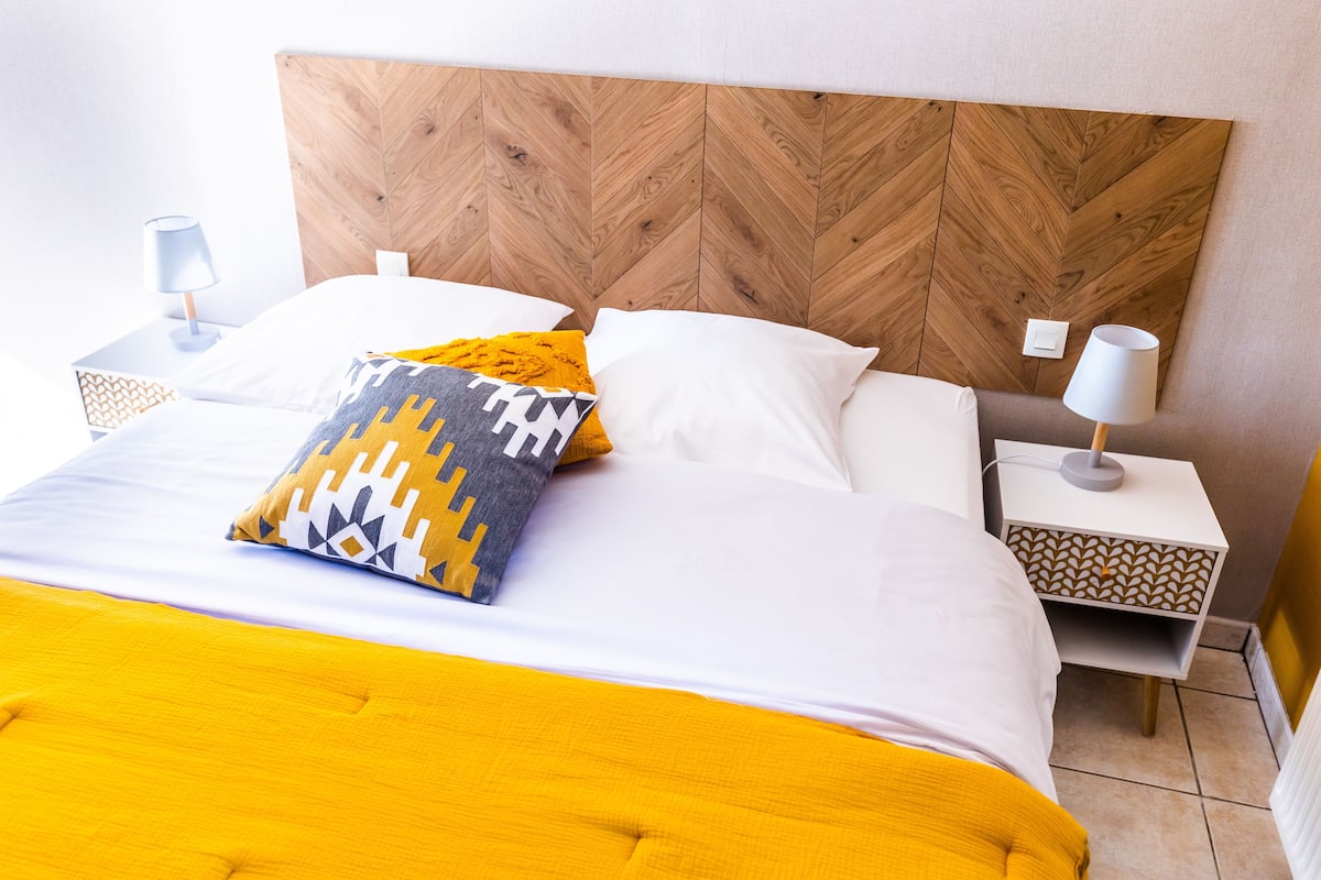 Cama bem arrumada com cabeceira de madeira, duas mesas de cabeceira com abajures e um cobertor estampado.