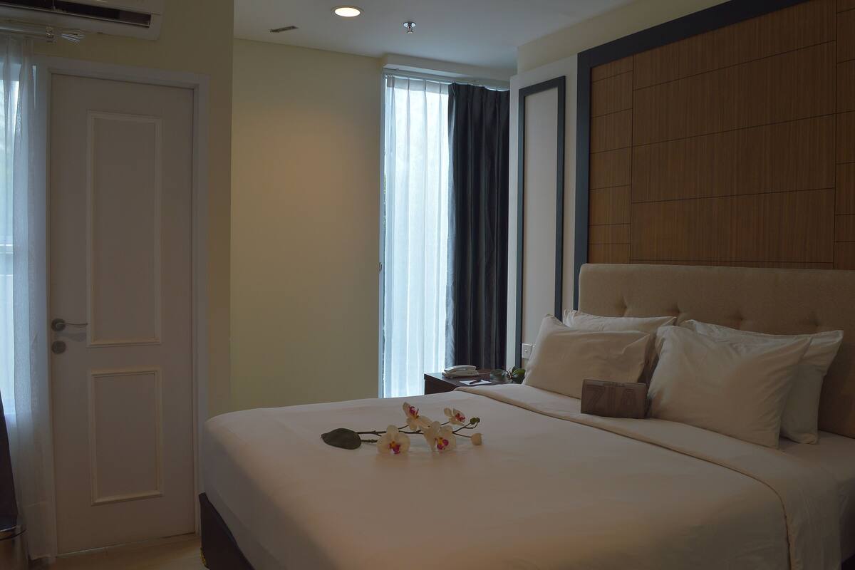 A hotel room with a large bed, a wooden headboard, a bedside table with a phone, and a window with curtains.