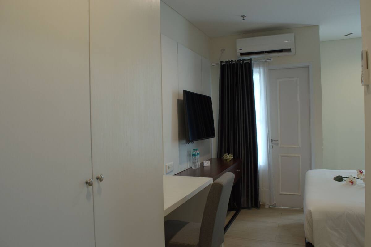 A hotel room with a bed, a desk with a chair, a television, and an air conditioning unit.