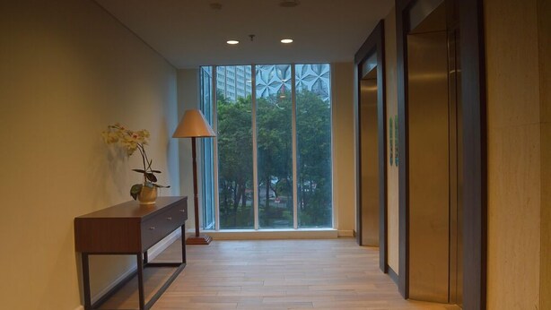 A hallway with a wooden floor, a desk with a vase of flowers, a lamp, and a large window with a view of trees and buildings. - Giia Sanno Menteng (Jakarta)